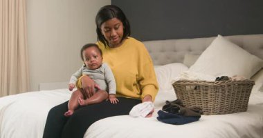 Genç bir anne çamaşır katlarken bebeği kucağında yatağında oturur. Giysi parçalarını alıyor ve hasır çamaşır sepetine koyuyor. Bu iç açıcı görüntü aile hayatının ve anneliğin özünü yansıtıyor..