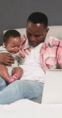 Bir baba bebeğiyle birlikte yatağında oturur ve uzaktan kumandayla çalışır. Bebek mızmızlanır ve ağlar. Babası onu kucağına alıp rahatlatıyor. Bu, evde çalışmanın ve ebeveynliğin zorluklarını yakalar..