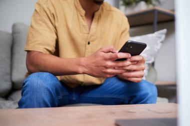 Kanepede oturan, elinde akıllı telefon tutan ve kullanan biri. Günlük kıyafet ve oturma odası modern bir ev ortamında rahatlık ve rahatlık hissi veriyor..