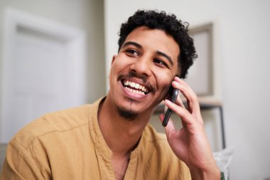 Neşeli bir adam kulağına cep telefonu dayamış içeride konuşurken neşeyle gülümsüyor. Sıcak ortam ve günlük giysiler rahat ve keyifli bir etkileşimi vurguluyor, dijital iletişimi en iyi şekilde gösteriyor.