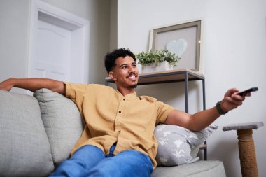 Gündelik bronz gömlekli bir adam, elinde uzaktan kumandayla koltukta rahatlar ve modern bir oturma odasında gülümser. Ortam hoş ve davetkar görünüyor, arka planda ince dekorlar var..