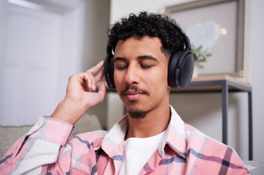 Genç bir adam evde rahatça oturup, ses geçirmez kulaklıklarla favori parçalarının tadını çıkarıyor. Huzurlu ifadesi, huzurlu bir ev ortamında rahatlamasını ve kaliteli sesten keyif almasını sağlıyor..