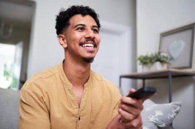Neşeli bir adam elinde uzaktan kumandayla parlak ve rahat bir oturma odasında oturuyor. Atmosfer sıcak ve davetkar. Bir anlık eğlence ve keyif sergiliyor..