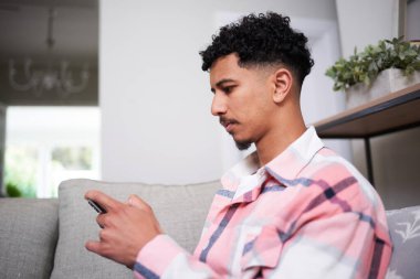 Genç bir adam rahat bir oturma odasında oturmuş, akıllı telefonunu rahatça kullanırken boş zamanlarının tadını çıkarıyor. Kapalı alan ortamında rahatlama ve modern yaşam tarzı aktiviteleri vurgulanıyor.