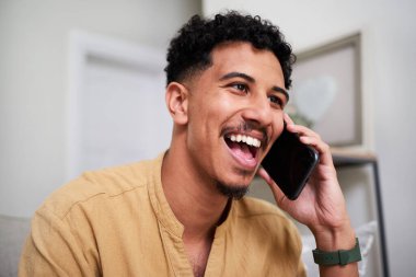 Neşeli bir adam gülümsüyor ve ev içi bir telefon görüşmesinden zevk alıyor, samimi bir neşe ve bağlantı anı yaşıyor. Sıcak atmosfer, modern bir ev ortamında arkadaşlığı, mutluluğu ve günlük bağlantıları vurguluyor..