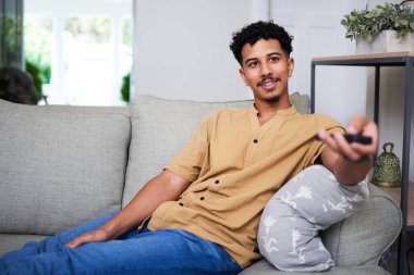 Sıradan giyimli bir insan, rahat bir oturma odasındaki modern bir kanepeye uzanır. Uzaktan kumandayla, rahat ve sade bir atmosfer yaratıyorlar..