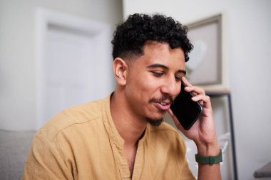 Evde rahat bir şekilde oturan ve cep telefonuyla konuşan genç bir adam. Doğal ışıklandırma konforlu ve rahat ortamı geliştirir, kişisel bağlantı ve iletişim anını yakalar..
