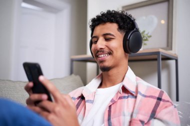 Neşeli genç bir adam evde boş zamanlarının tadını çıkarıyor, rahatça oturuyor ve akıllı telefonuna bakarken kulaklık takıyor. Rahat, sakin kapalı atmosfer rahatlamayı ve bağlantıyı vurguluyor..