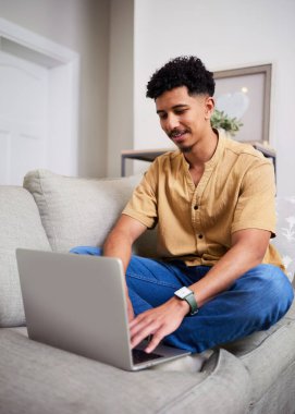 Bronz gömlek ve kot pantolon giymiş genç bir adam dizüstü bilgisayarıyla çalışırken ışık dolu bir oturma odasında kanepede bacak bacak bacak üstüne atmış oturuyor. Sahne rahat ve üretken bir atmosfer sunuyor..