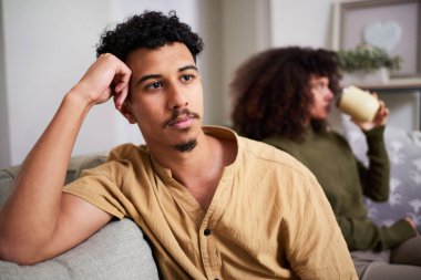 Genç bir adam düşünceli bir ifadeyle bir kanepede oturuyor, başını elinin üzerine yaslıyor. Arka planda, başka bir kişi rahat bir evde içki içerken rahatlıyor..