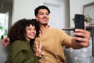 Neşeli bir çift, rahat bir oturma odasındaki kanepede oturup gülümseyen ve barış işareti olan bir selfie için poz veriyor. Çevre sıcaklığı ve gevşemiş, neşeli bir atmosferi gösteriyor..