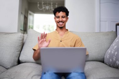 Neşeli bir adam modern bir oturma odasında rahat bir şekilde oturur ve bilgisayarla sanal bir toplantı yapar. İçten bir şekilde gülümser ve ekranda el sallar..