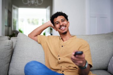 Neşeli genç bir adam rahat bir koltukta oturuyor, elinde uzaktan kumandayla televizyon seyrediyor ve rahat bir atmosferin tadını çıkarıyor..