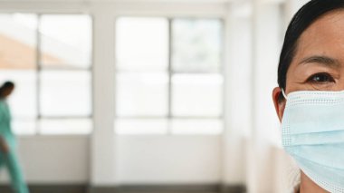 Koruyucu maske takan bir sağlık uzmanının odaklanmış görüntüsü. Önlüklü başka bir figürle parlak atmosfer, tıbbi ortamlarda profesyonel bakım ve güvenliği vurguluyor..