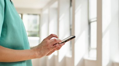 Önlük giyen bir sağlık çalışanı, modern pencerelerden ışık saçan parlak bir çevrede dururken akıllı telefon kullanmakla meşgul. Sağlık hizmetlerindeki teknolojiyi temsil ediyor.