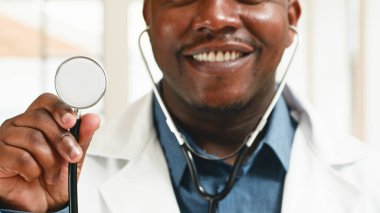 Beyaz önlük giyen ve elinde steteskop tutan bir sağlık görevlisi evde güvenle gülümsüyor. Bu görüntü, güven ve iletişimi desteklemek için ideal bir sağlık ortamında profesyonellik, adanmışlık ve yaklaşımı iletir..