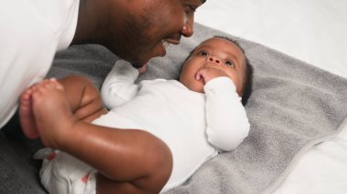 Bir baba bebeğiyle sevgi dolu bir şekilde etkileşime girer, sıcak ve besleyici bir an yaratır. Bebek gri bir havluda yatarken baba bebeğe gülümsüyor ve pozitif bir bağ kuruyor..