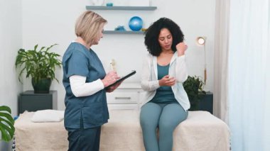 Kıdemli bir fizyoterapist, muhtemelen küçük bir muayenehanenin sahibi genç bir siyahi müşteriyle detaylı bir istişarede bulunuyor. Müşteri, spor fizyolojisi veya iyileşme için masaj konusunda uzman tavsiyesi isterken dirsek ağrısını tarif ediyor.. 