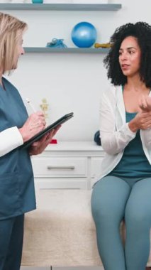 Deneyimli bir kıdemli fizyoterapist bir tablete not alırken genç bir siyahi kadın hasta bilek ağrısını tarif ediyor ve hareketlerini gösteriyor. Bu profesyonel konsültasyon rehabilitasyon ve yaraların iyileşmesine odaklanmaktadır..