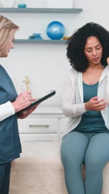 Deneyimli bir kıdemli fizyoterapist, omuz ağrısı ve yaralanmayı tanımlayan siyah bir kadın müşteriye özenle danışıyor. Sağlık uzmanı, klinikte uzman değerlendirmesi ve bakım sunan bir tablete dikkatle not alır..