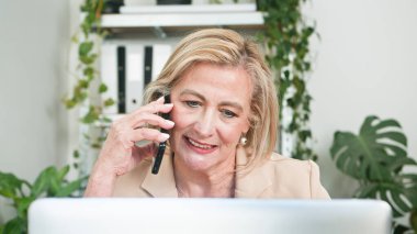 Yaşlı profesyonel bir kadın birden fazla iş yapıyor, dizüstü bilgisayarla çalışıyor ve yeşil, bitkilerle dolu bir ofis ortamında telefonla konuşuyor..