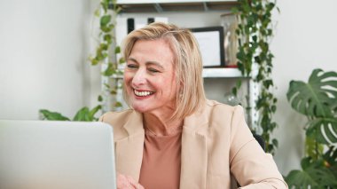 Sanal bir toplantıya katılırken gülümseyen yaşlı profesyonel bir kadın. Sahne, iç mekan bitkileriyle süslenmiş bir iş ortamında işbirliği, uzmanlık ve yaratıcılık taşıyor..