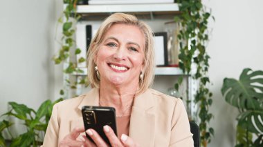 Üst düzey bir kadın evde akıllı bir telefonu tutarken gülümsüyor, iş ortamında bitkilerle çevrili, profesyonellik ve bağlantı duygusunu iletiyor.