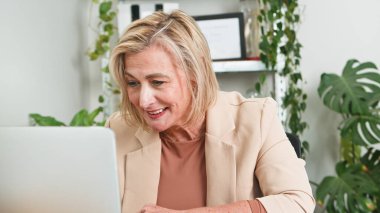 İş kıyafeti giyen daha yaşlı profesyonel bir kadın, modern ve bitki desenli bir ofiste dizüstü bilgisayarda oturan işine odaklanmış durumda..