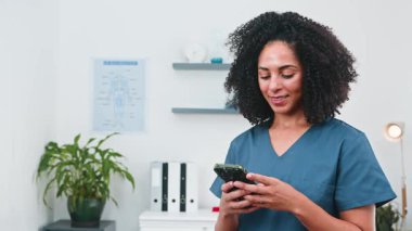 Kendine güvenen, gülümseyen ve kameraya bakan farklı kadın fizyoterapist profesyonel modern kliniğinde akıllı telefonunu kullanıyor. Sağlık, spor bilimi ve başarılı bir küçük işletmeyi temsil ediyor..