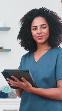 Önlüklü profesyonel kadın fizyoterapist elinde dijital tablet tutarken güvenle gülümsüyor. Modern sağlık hizmetlerini, tıbbi uzmanlığı ve teknolojinin hasta bakımına entegrasyonunu, çeşitliliği ve spor bilimi bilgisini temsil ediyor..