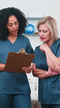 İki profesyonel kadın fizyoterapist hasta bakımı ve tedavi planlarını tartışan bir panoda tıbbi raporları gözden geçiriyorlar. Onların işbirliği çalışmaları klinik ortamında sağlık ve spor bilimine olan bağlılıklarını gösteriyor..