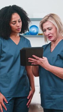 Kendini adamış iki kadın fizyoterapist hasta bilgilerini dijital tablette gözden geçiriyorlar. Spor bilimi uygulamalarını tartışıyor ve modern bir klinikte çeşitli profesyonel sağlık uzmanlıklarının altını çizerek tedavi planları üzerinde işbirliği yapıyorlar..