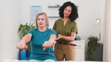 Deneyimli bir siyahi kadın fizyoterapist, yaşlı bir hastaya klinik ortamında halterlerle rehabilitasyon uygulamasında yardımcı oluyor. Spor bilimi prensiplerini vurgulayan bu oturum, sağlık ve iyileşmeyi teşvik etmektedir..