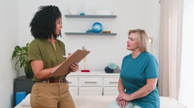 Şefkatli siyah bir kadın fizyoterapist, boyun ve omuz rahatsızlığı olan yaşlı bir kadın hastaya danışmanlık yapıyor. Modern bir klinikte kişisel bakım ve iyileşme desteği sağlayarak belirtileri not alıyor..