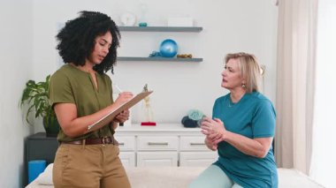 Profesyonel bir siyahi fizyoterapist yaşlı bir kadın hastaya danışmanlık yapar, sağlık sorunlarını anlamak ve iyileşme ve iyileşme için etkili bir rehabilitasyon planı geliştirmek için özenle not alır..