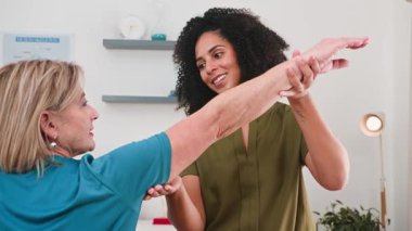 Şefkatli bir siyahi kadın fizyoterapist, kollarını iyileştirme egzersizlerinde ona rehberlik eden yaşlı bir kadın hastayla özenle çalışıyor. Bu profesyonel etkileşim modern bir klinik ortamında bakım, iyileşme ve hareket kabiliyetinin geliştirildiğini vurgulamaktadır..