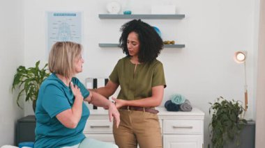 Şefkatli bir siyahi kadın fizyoterapist yaşlı bir hastaya hedef alınan egzersizlerle kol hareketlerini geliştirmesinde yardımcı olur. Bu profesyonel rehabilitasyon oturumunda hareketin düzeltilmesi ve genel sağlığın iyileştirilmesi üzerinde duruluyor..