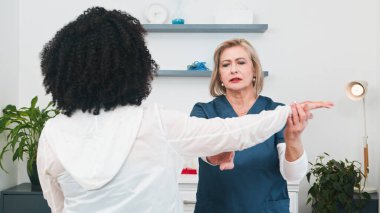 Üst düzey bir fizyoterapist, küçük bir ağırlık kullanarak hedef alınan omuz rehabilitasyonu egzersizlerinde genç bir siyahi müşteriye dikkatle yardım eder. Bu odaklanmış fizik tedavi seansı parlak, modern bir özel muayenehane içinde gerçekleşir.