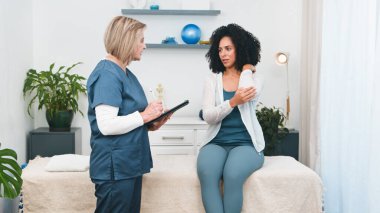 Kıdemli bir fizyoterapist, muhtemelen küçük bir muayenehanenin sahibi genç bir siyahi müşteriyle detaylı bir istişarede bulunuyor. Müşteri, spor fizyolojisi veya iyileşme için masaj konusunda uzman tavsiyesi isterken dirsek ağrısını tarif ediyor.. 