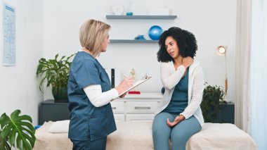 Yetenekli bir kıdemli fizyoterapist genç bir siyahi bayan müşteriyle kapsamlı bir istişarede bulunuyor. Müşteri boynunu ve omzunu işaret ediyor.. 