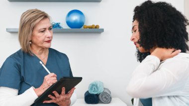 Şefkatli bir kıdemli fizyoterapist detaylı bir muayene sırasında genç bir siyahi kadın müşterinin boyun ağrısı ve sağlık sorunlarını özenle kaydeder..