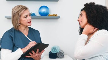 Üst düzey bir fizyoterapist genç bir siyahi müşteriye boyun ağrısı ve olası sakatlığı hakkında dikkatle danışıyor. Tecrübeli spor fizyoterapisti dijital tablete notlar alır, bu da küçük muayenehanesinde profesyonel bakımı gösterir.. 