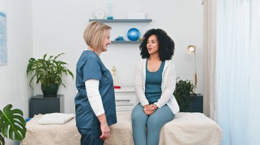 Deneyimli bir kıdemli fizyoterapist genç siyah bayan müşterisiyle sıcak ve profesyonel bir istişare içine girer. Sağlık hizmetleri ve küçük işletme temaları için ideal.