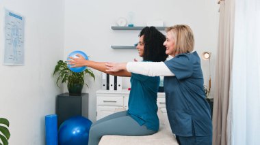 Deneyimli bir kıdemli fizyoterapist titizlikle genç bir Afrikalı Amerikalı kadına küçük bir top ile doğru duruş ve hareketi vurgulayan hassas bir fizik tedavi egzersizinde rehberlik ediyor.. 