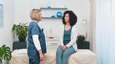 Üst düzey bir fizyoterapist modern özel muayenehanesinde genç siyahi müşterisini sıcak bir şekilde selamlıyor. Spor fizyoterapisi veya genel rehabilitasyon için bir danışma, daha rahat bir ortamda kişisel sağlık hizmetlerinin vurgulanması.