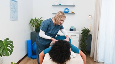 Deneyimli bir kıdemli fizyoterapist genç siyahi müşterilerine özel spor masajı yapar. Kas iyileşmesini ve ağrı kesiciyi teşvik eder. Bu profesyonel manuel terapi seansı refahı ve uzman bakımını ön plana çıkarıyor.