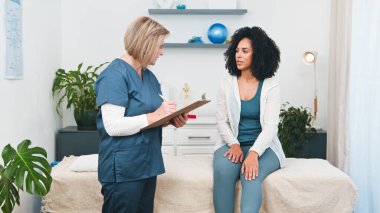Üst düzey bir fizyoterapist, profesyonel, küçük bir klinikte genç bir siyahi müşteriyle uzmanca sağlık ve rehabilitasyon konularını tartışır. Modern bir sağlık ortamında iyileşme ve genel refah için şefkatli bir yaklaşım..