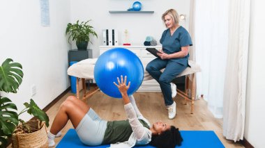 Kendini adamış kıdemli bir fizyoterapist genç bir siyahi müşteri için kapsamlı spor fizyoterapisi ve rehabilitasyon sunuyor. Egzersiz topuyla müşteriye ustalıkla rehberlik eder, iyileşme ve refahı destekler..