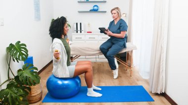 Deneyimli bir kıdemli fizyoterapist genç bir Siyahi müşteriyi özenle gözlemler ve fizik tedavi sırasında rehberlik eder. Müşteri egzersiz topunun üzerinde otururken halterlerle güç egzersizleri yapar ve rehabilitasyona odaklanır.