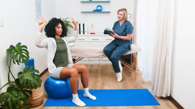 Üst düzey bir fizyoterapist genç bir siyahi müşteriye egzersiz topunun üzerindeki dambıllarla egzersizleri güçlendirerek rehberlik eder. Küçük, profesyonel bir fizik tedavi uygulamasında bu seans uzman bakımı ve kişisel rehabilitasyonu vurguluyor..